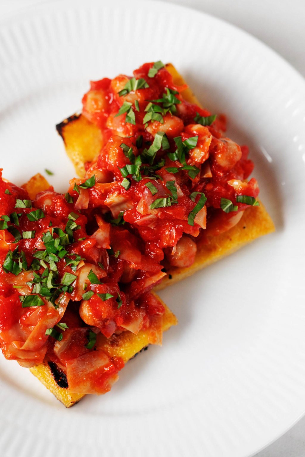 An overhead image of two slices of grilled polenta, which rest on a white dinner plate. They're smothered in a tomato sauce and chickpea mixture.