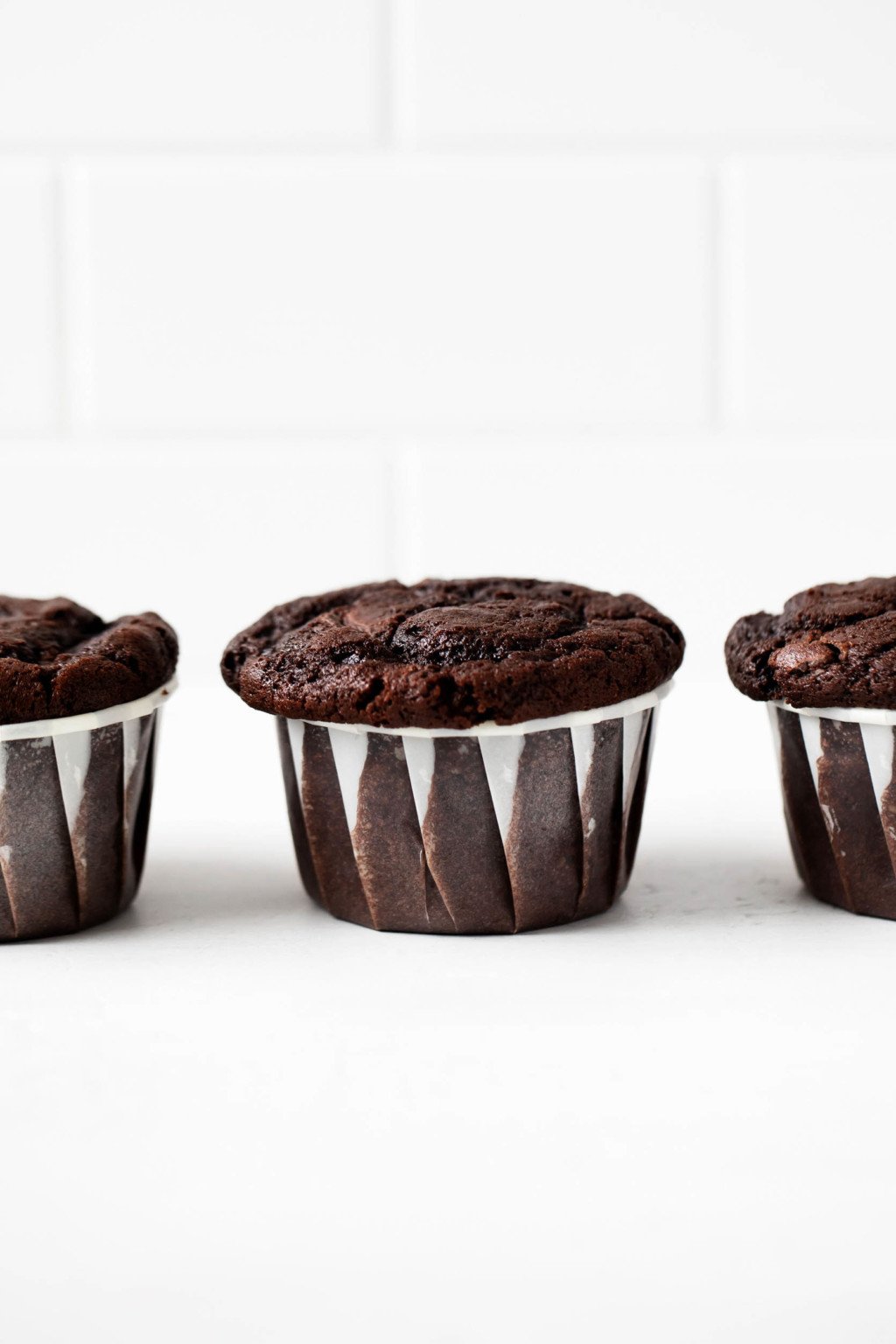 A few vegan double chocolate muffins have been lined up in a row, right in front of a white brick surface.