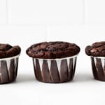 A few vegan double chocolate muffins have been lined up in a row, right in front of a white brick surface.