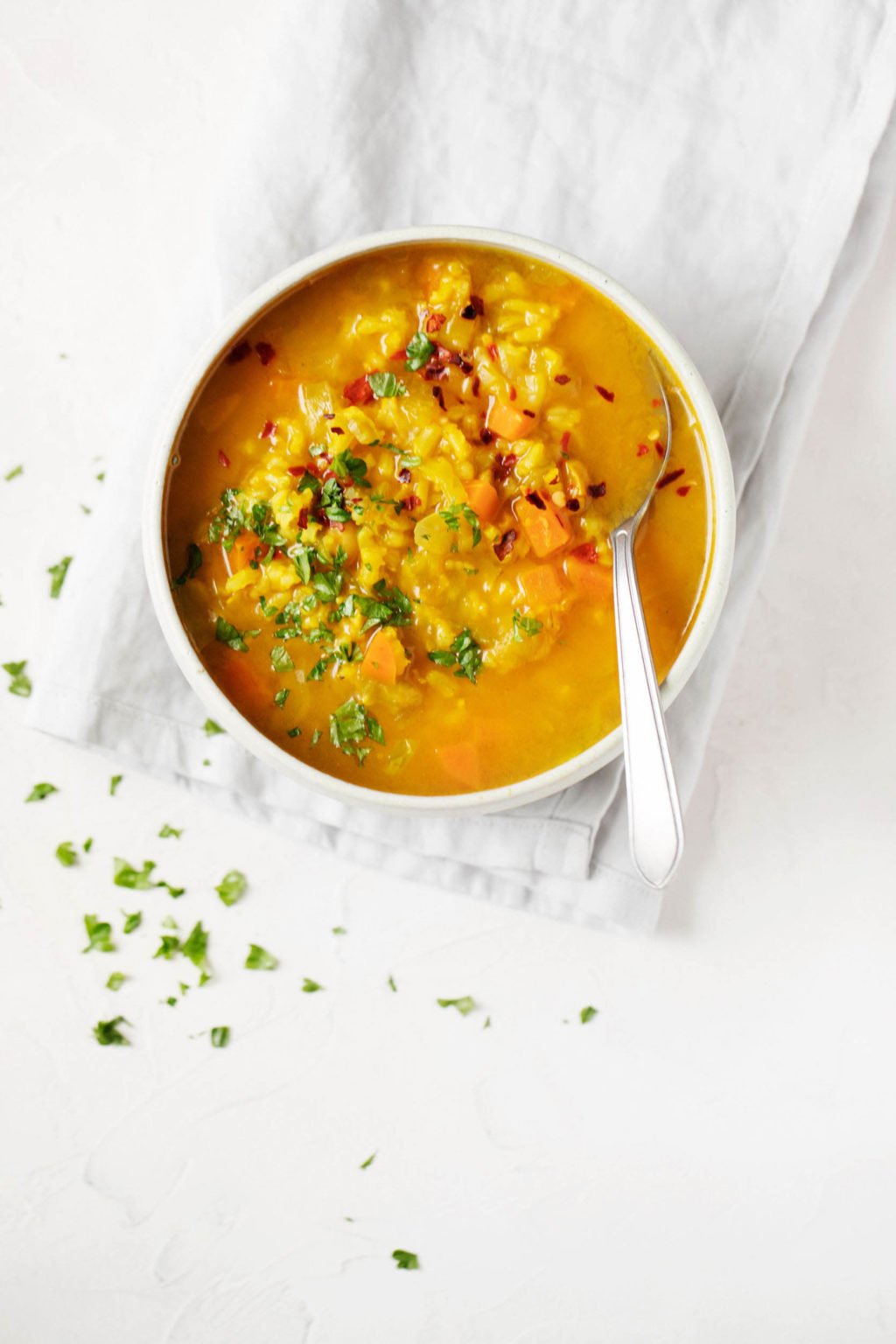 A folded gray napkin is being covered by a golden hued bowl of soup. There are specks of green parsley sprinkled around the napkin. 