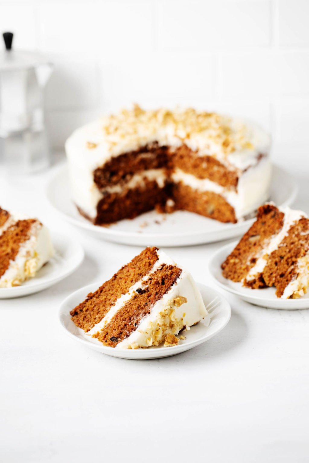 A vegan layered carrot cake is resting on a round platter. It has been sliced into three slices, which are each resting on small dessert plates against a white surface.