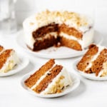 A vegan layered carrot cake is resting on a round platter. It has been sliced into three slices, which are each resting on small dessert plates against a white surface.