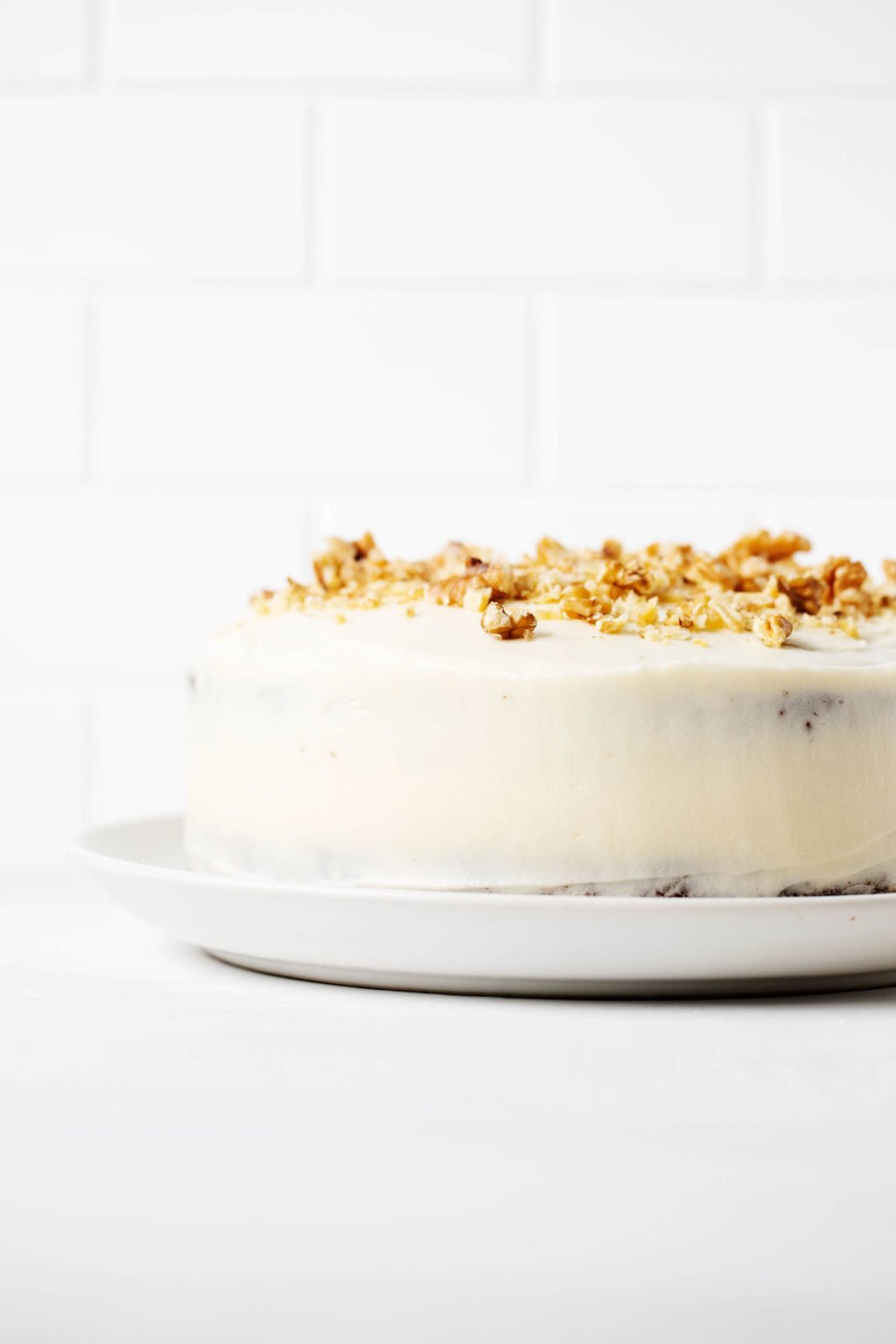 An angled photograph of a cake, which has been decorated with white frosting and chopped walnuts. It's resting on a white surface and against a white brick backdrop.