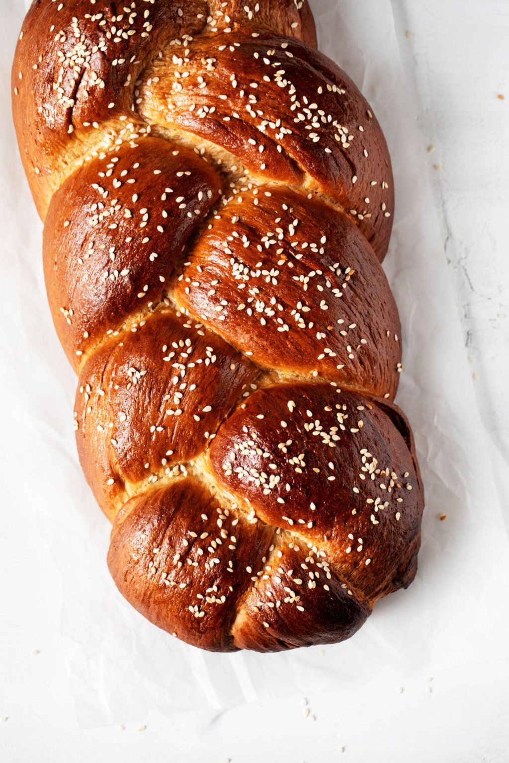 A braided plait of a three strand loaf of bread, garnished with sesame seeds and resting on white parchment.