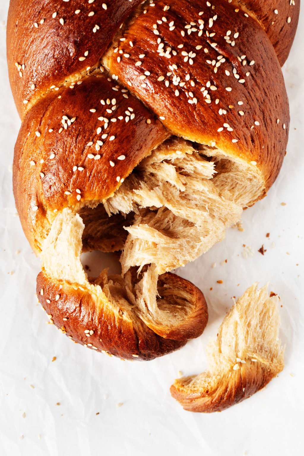 An overhead photo of torn, tender vegan Tsoureki bread, which is decorated with sesame seeds and resting on white parchment.