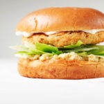 An angled photograph of a vegan artichoke white bean burger, served on a bun with greens and a creamy dip.