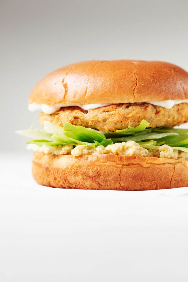 An angled photograph of a vegan artichoke white bean burger, served on a bun with greens and a creamy dip.