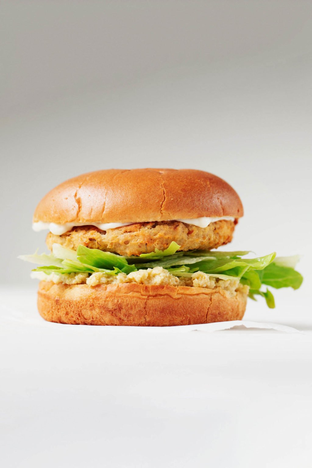 A sideways photograph of a vegetable burger on a bun, with lettuce and other toppings. It's on a white surface and photographed against a gray backdrop.