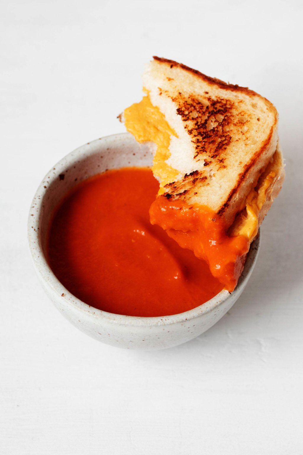 A small, white bowl of tomato soup has been dipped into with a half of a vegan grilled cheese sandwich.