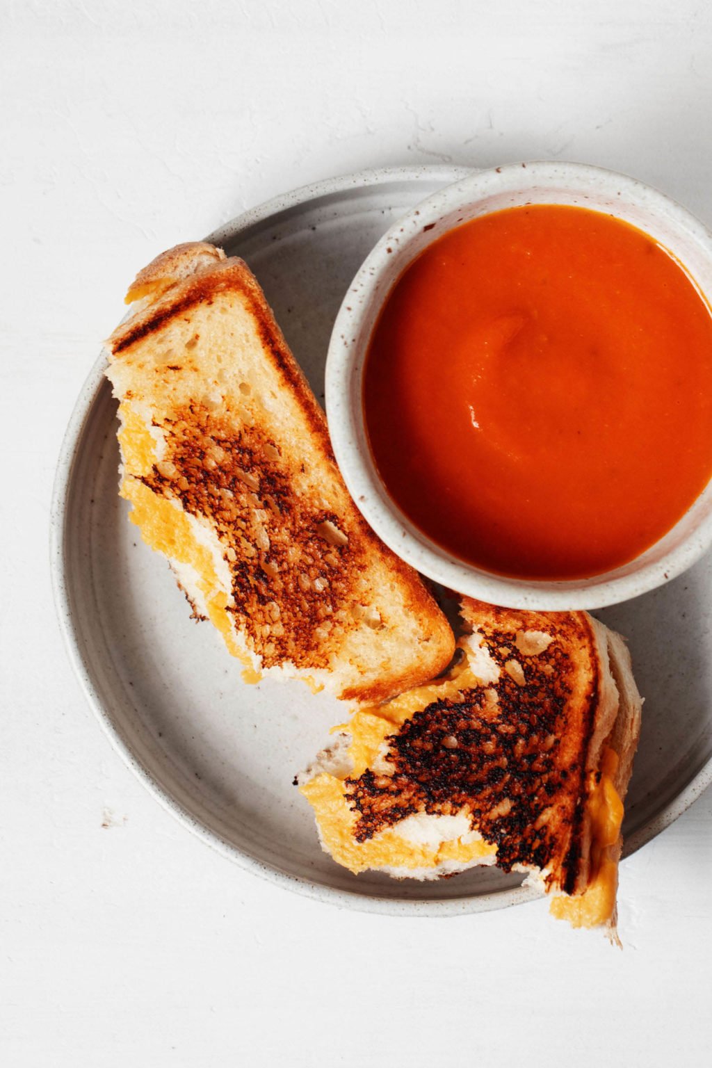 A small bowl of tomato soup is accompanied by a crispy vegan grilled cheese. Both rest on a small, round white plate.