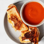 A small bowl of tomato soup is accompanied by a crispy vegan grilled cheese. Both rest on a small, round white plate.