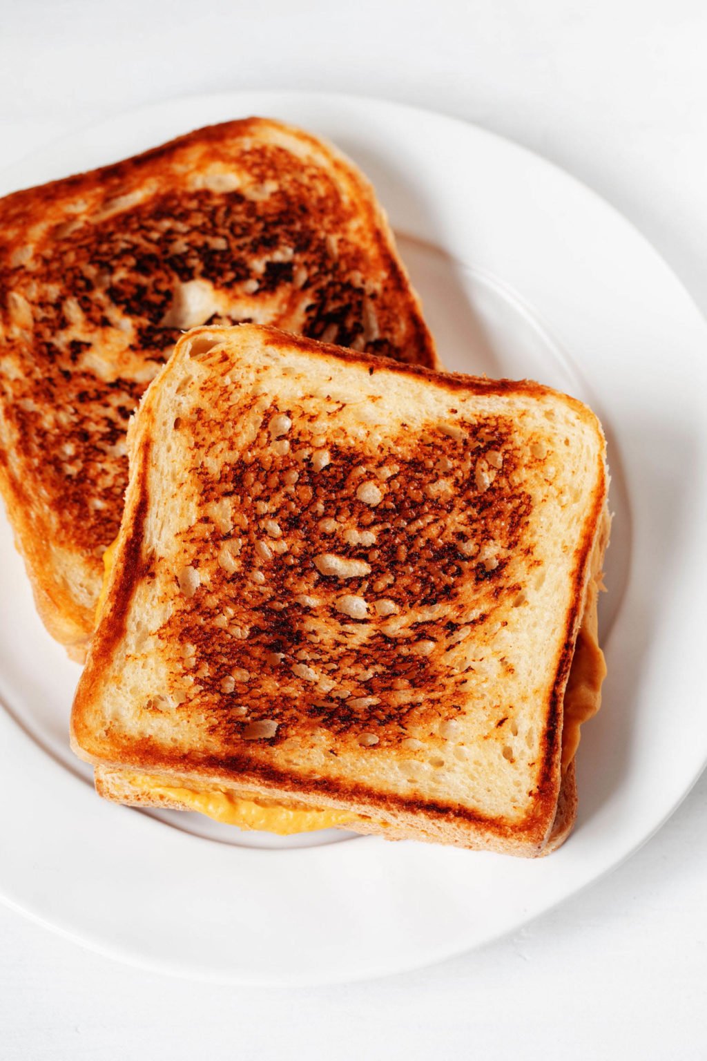 A toasted vegan grilled cheese sandwich is resting on a rimmed, white plate. The plate is resting on a white surface.