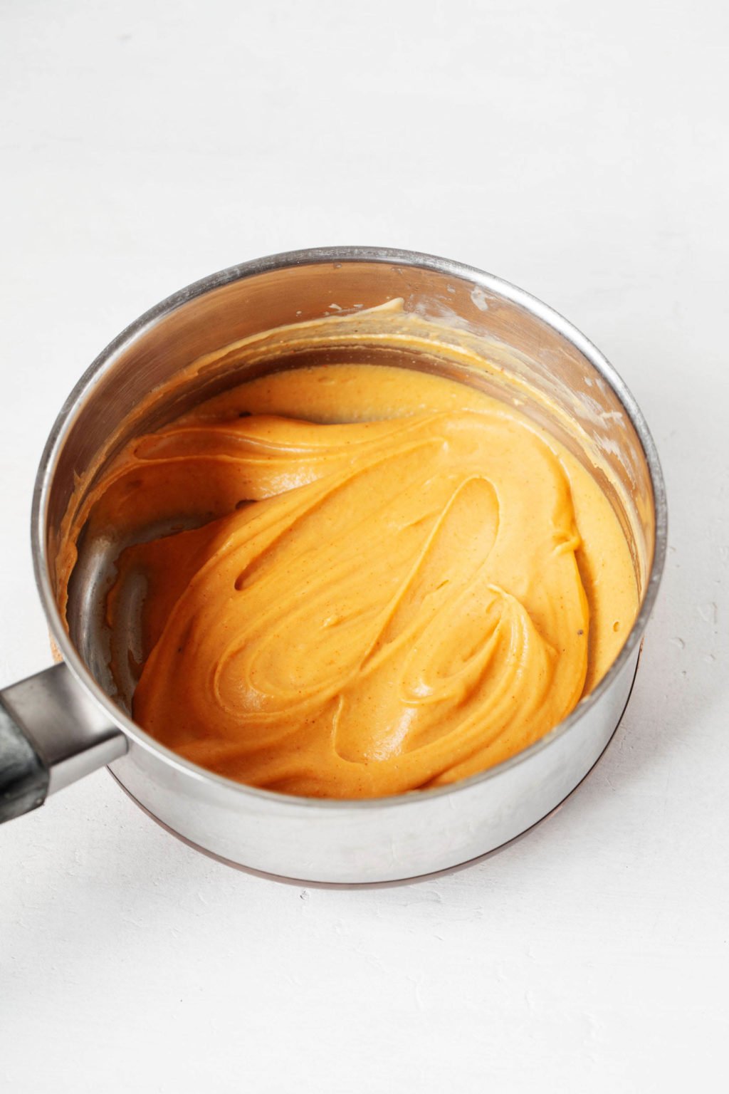 A silver saucepan rests on a white surface. It's filled with a light orange, thick sauce for vegan grilled cheese sandwiches.