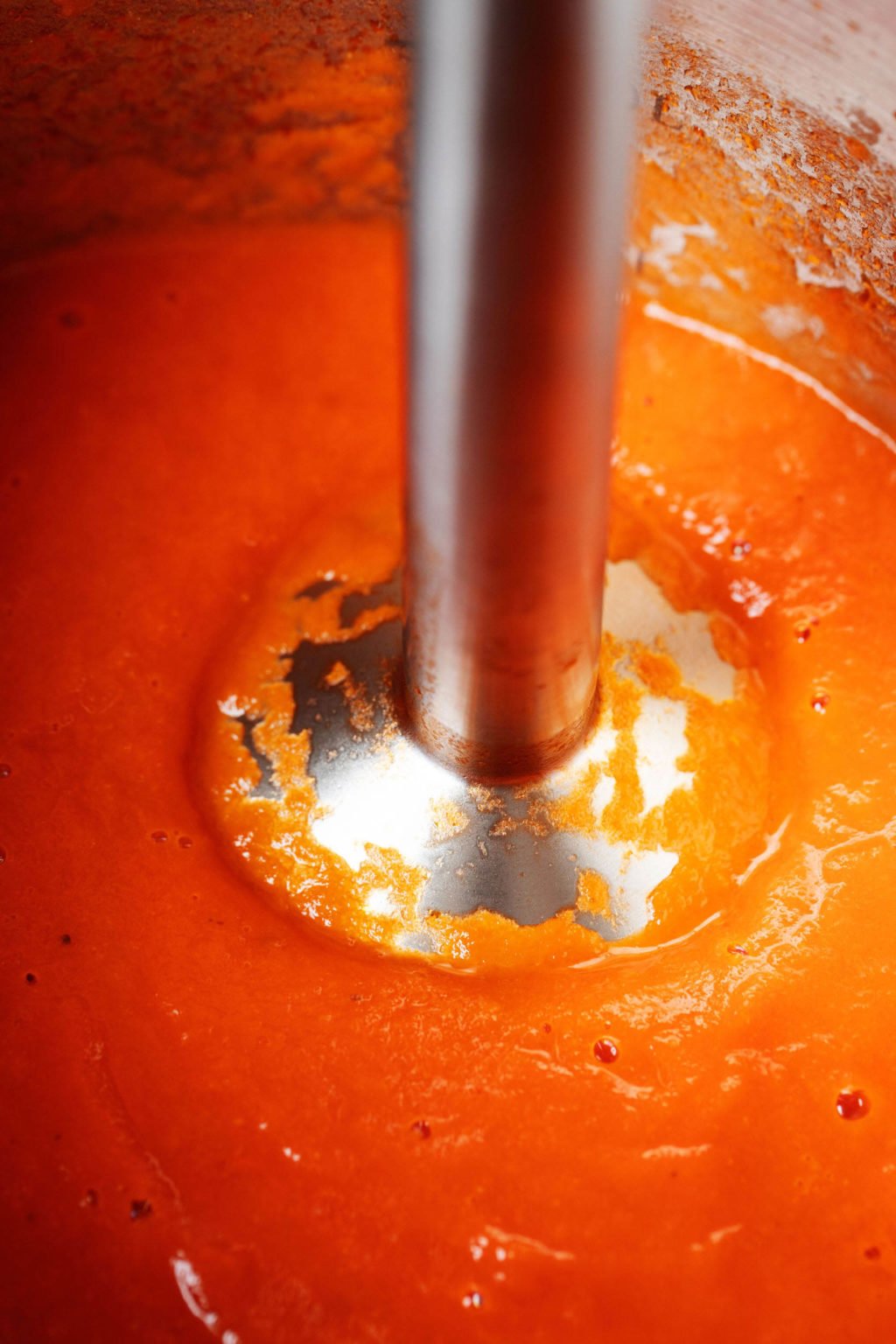 An immersion blender is submerged into a pot of tomato soup.