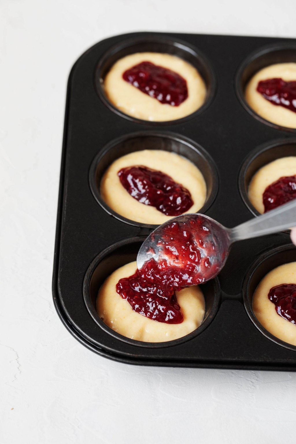 Corn muffins rest in a muffin pan. They're being filled with dollops of red raspberry jam.