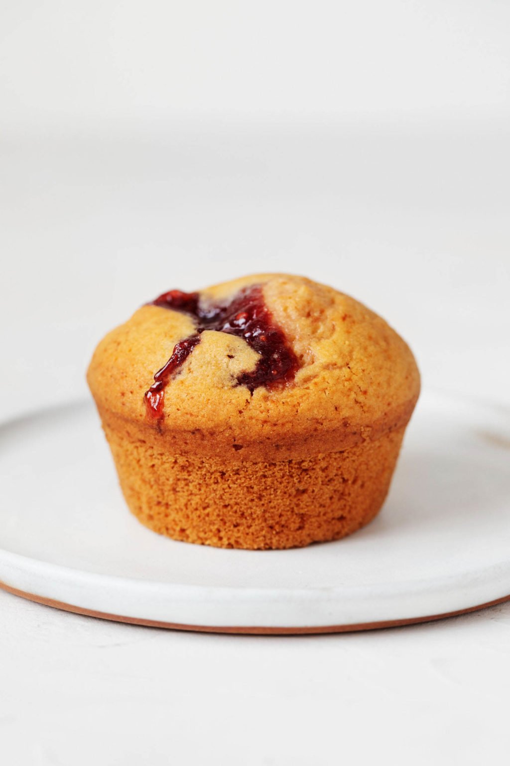 A vegan baked good with raspberry preserves is resting on a small white dessert plate. There is a white background behind it.