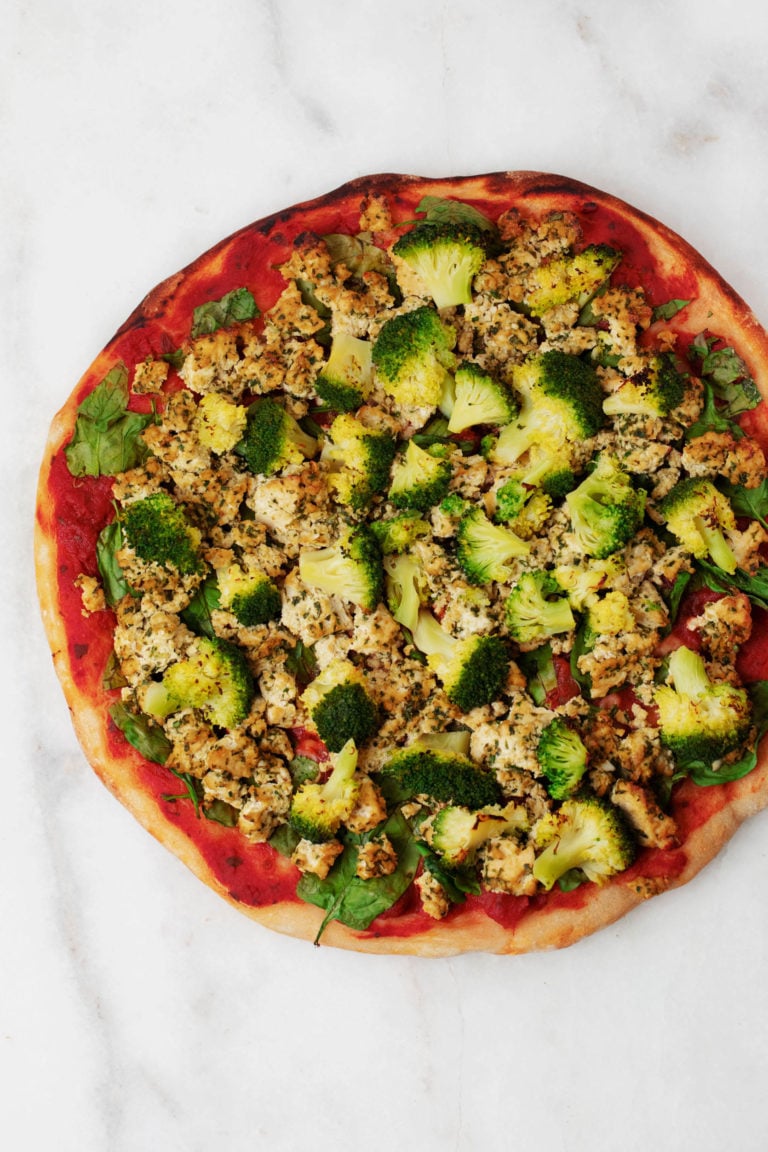 A vegan pizza made with herbed tofu and green vegetables rests on a marble surface.