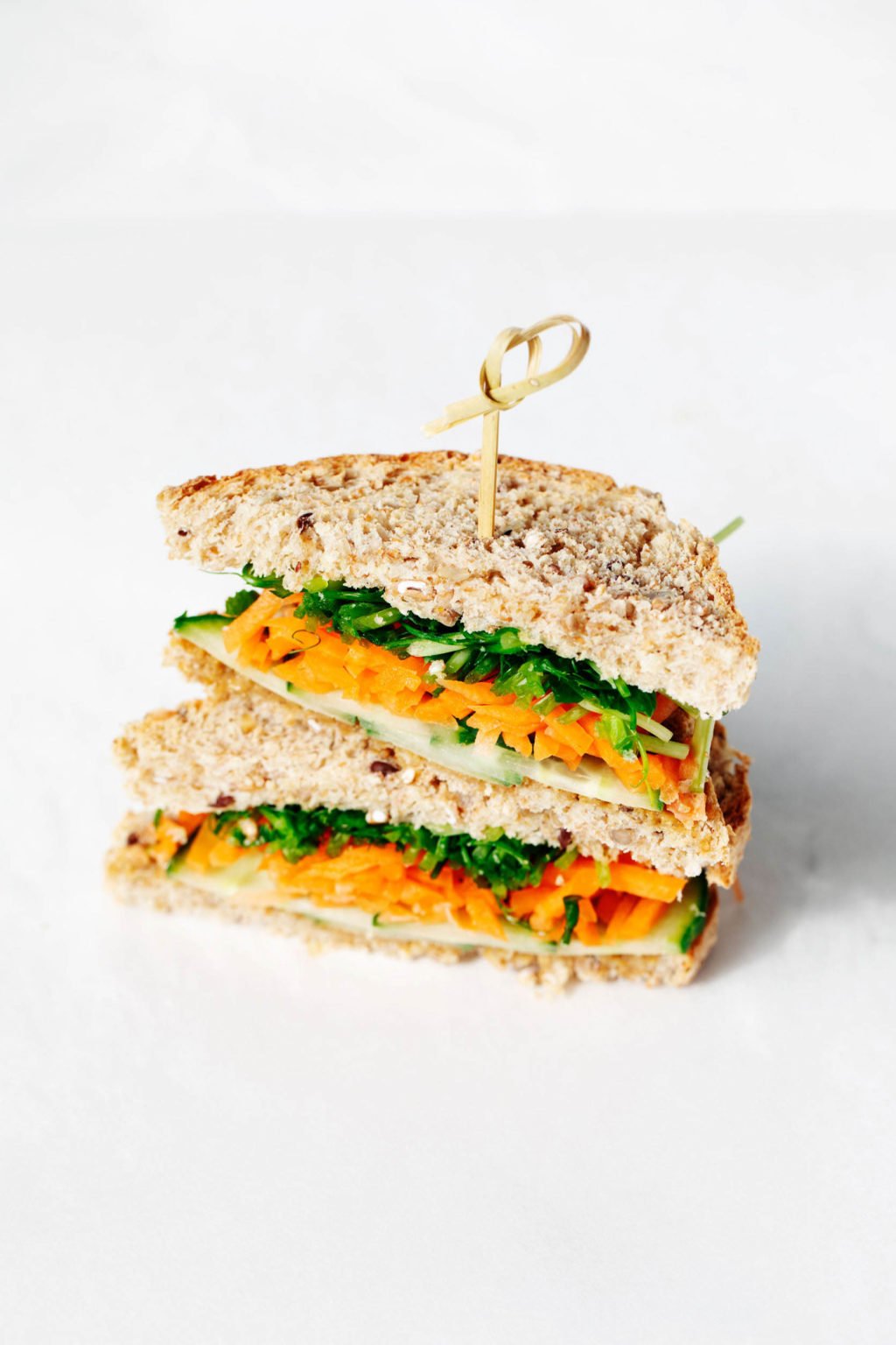 A whole grain, vegan miso tahini sandwich with fresh vegetables is stacked on a white surface.