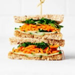 A sandwich has been assembled with layers of carrot, sprouts, and cucumber slices. It's resting on a white surface against a white brick backdrop.