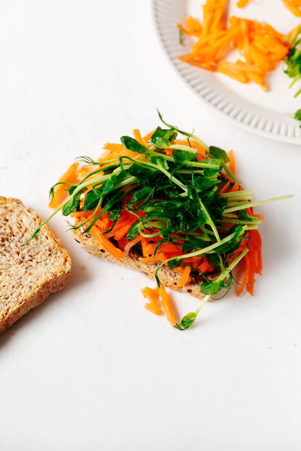 Two slices of whole grain bread are on a white surface. One slice has been piled with carrots and sprouts.