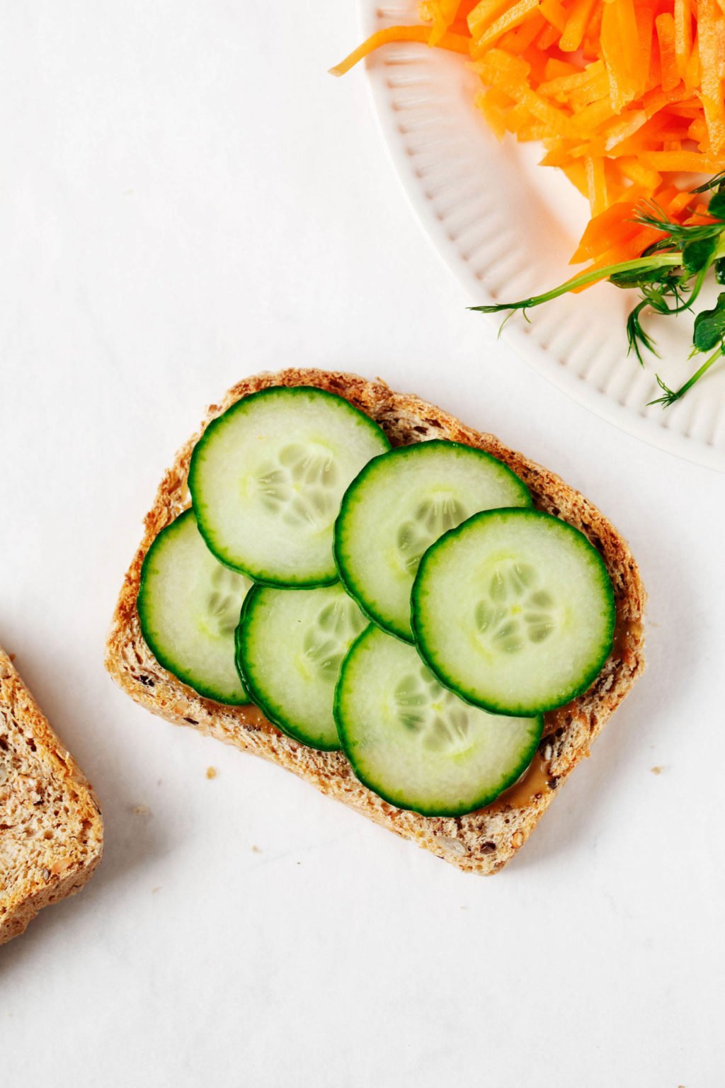 Two slices of whole grain sandwich bread are being assembled into a vegetable sandwich. They rest on a white surface.