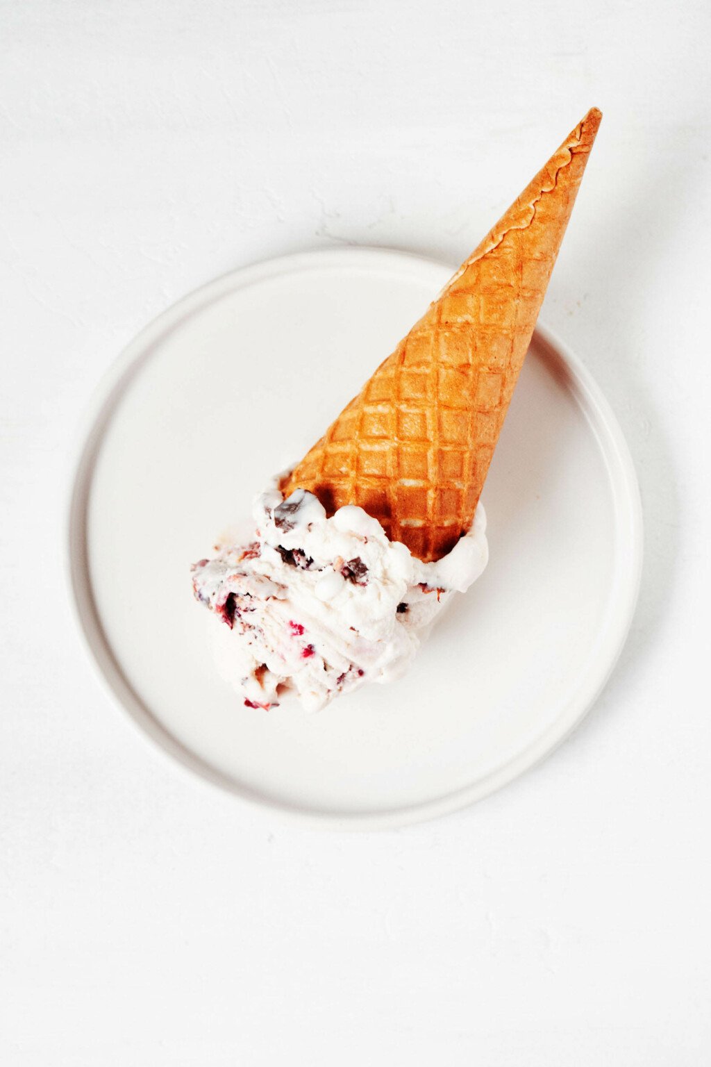 A small, round white plate holds an ice cream sugar cone with a scoop of cherry ice cream.