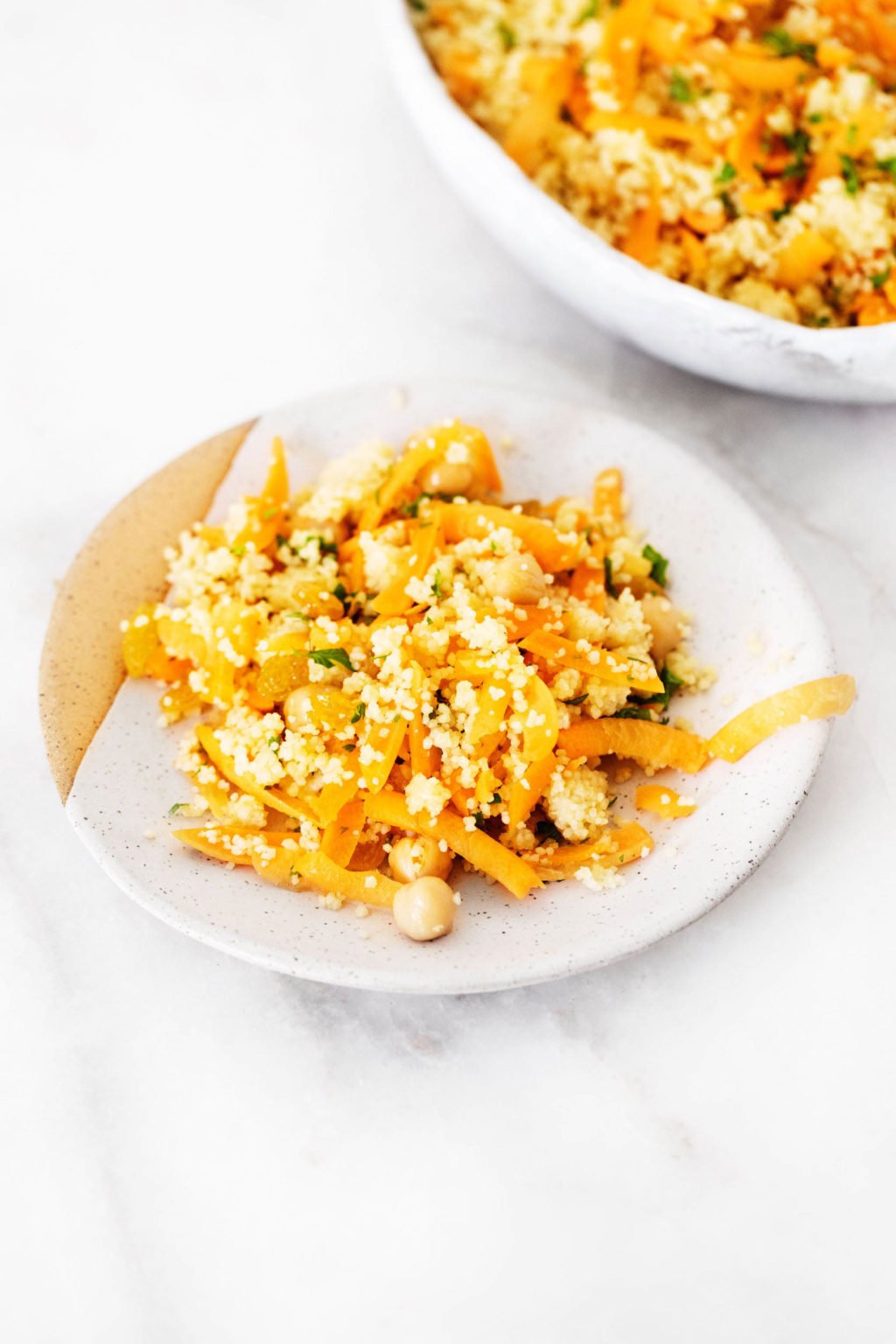 Two small plates serve a couscous salad that includes legumes and fresh herbs. They're resting on a white marble surface.