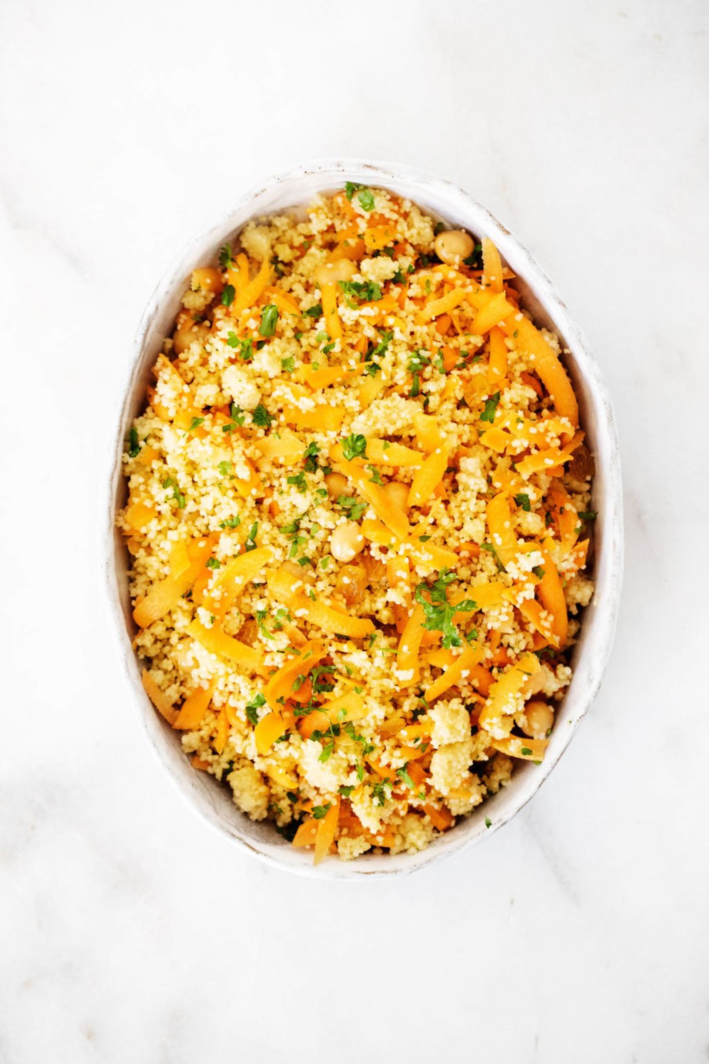 An oval shaped, white serving dish is filled with a couscous and chickpea salad, made with herbs and carrot. It rests on white marble.