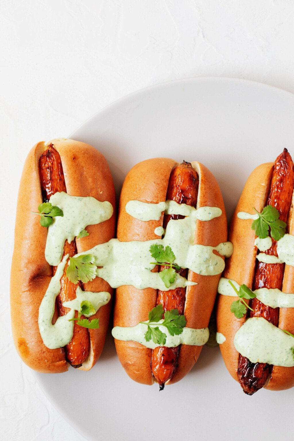 Carrot dogs, topped with a bright green sauce and chopped fresh herbs, are arranged on a round, white plate.