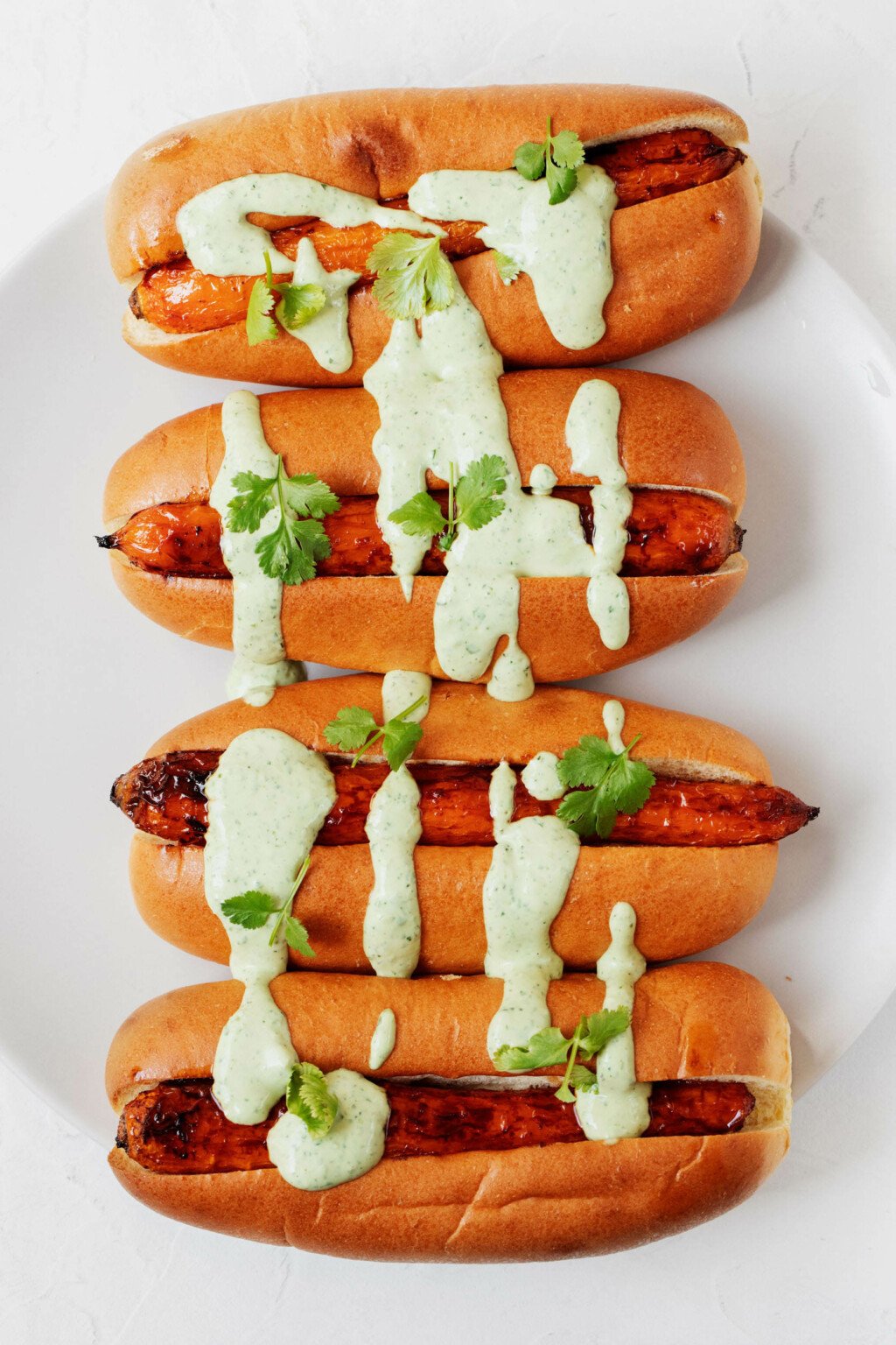 A white plate holds roasted carrots that have been arranged in hot dog buns and topped with a green sauce.