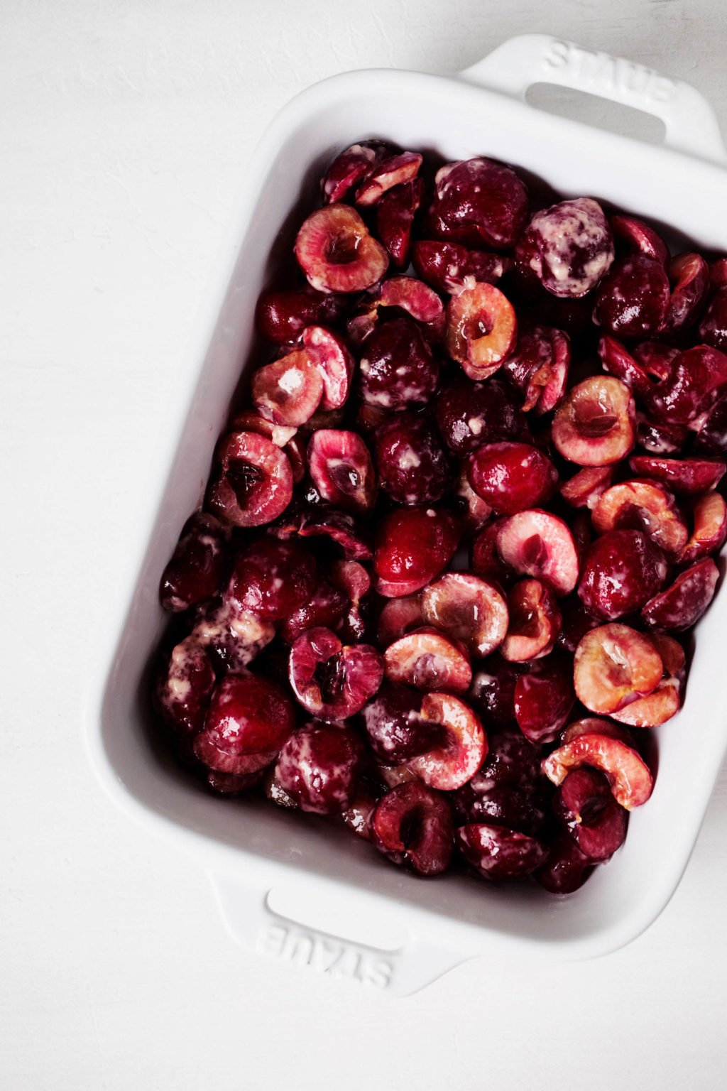 A white, rectangular baking dish is filled with deep red cherries that have been coated with sugar.