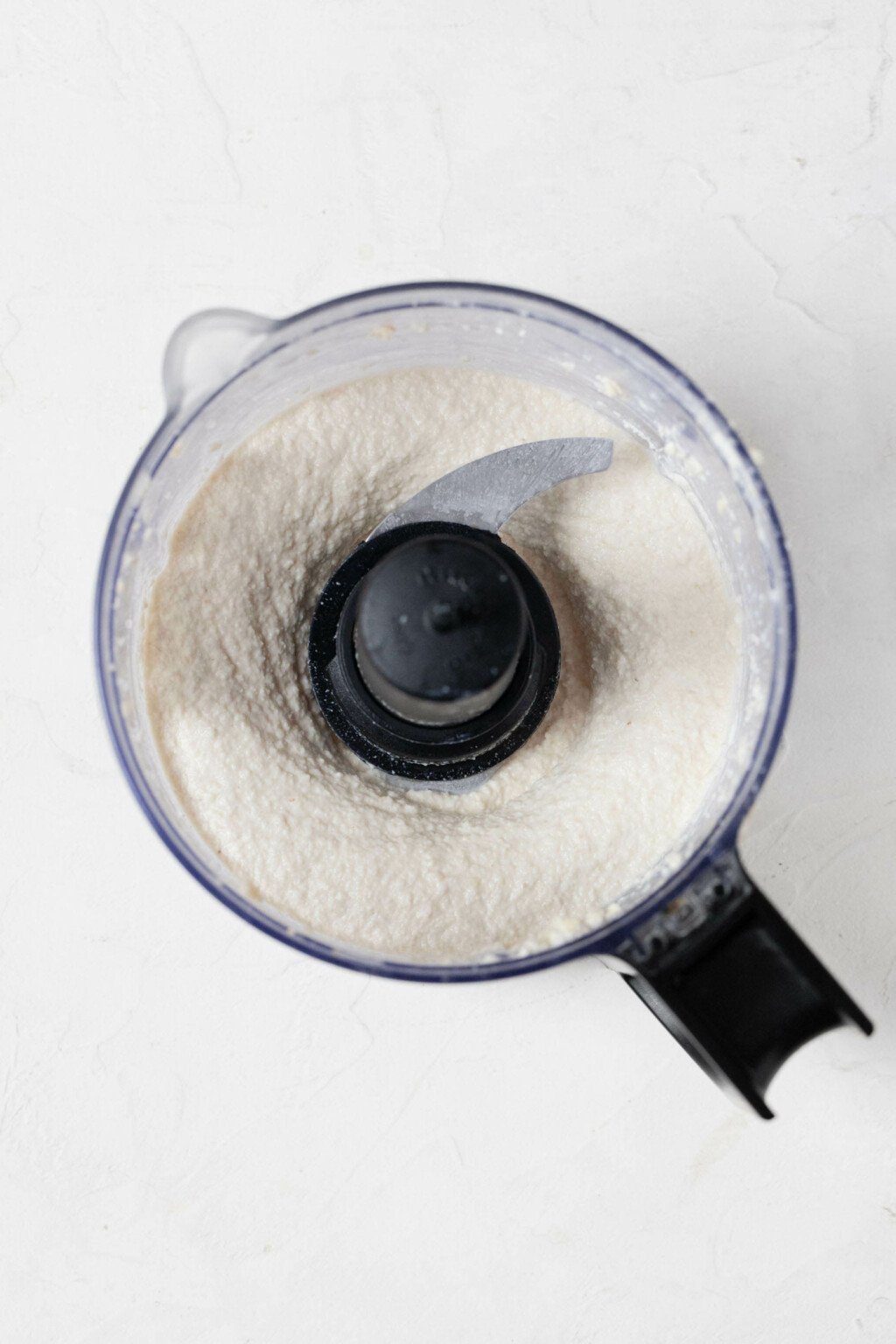 An overhead image of the bowl of a food processor, which is being used to prepare a creamy mixture with cashews and vegan cream cheese.