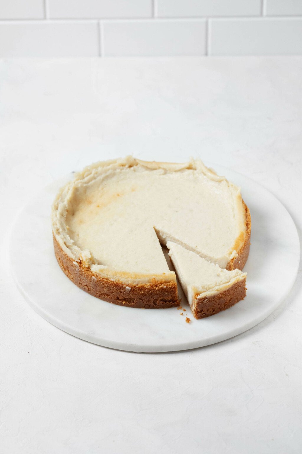 A freshly baked vegan cheesecake is positioned on a round, white serving dish. A slice has just been cut from the cake. 