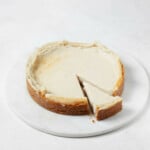 A freshly baked vegan cheesecake is positioned on a round, white serving dish. A slice has just been cut from the cake.