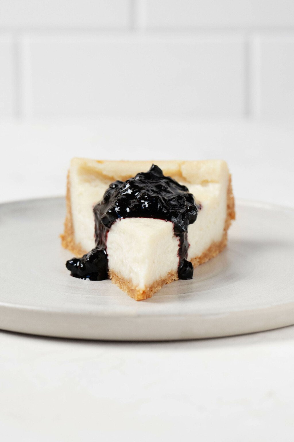 A slice of ultra creamy vegan cheesecake has been cut and topped with a dollop of blueberry sauce. It rests on a round, ceramic plate.