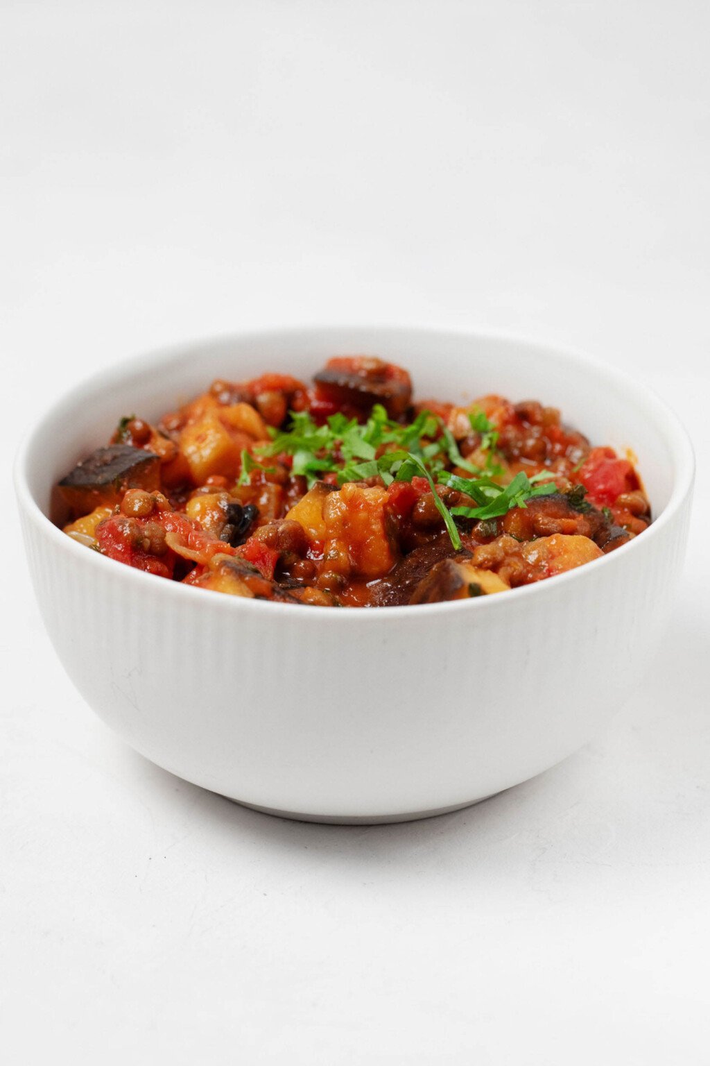 An image of stewed tomatoes, eggplant, and lentils. They're served in a while, round bowl and topped with chiffonade of fresh green herbs.