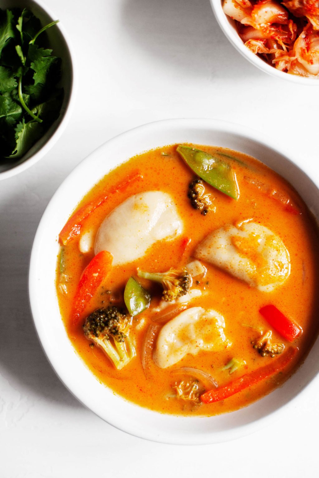 An overhead photograph of a warm bowl of red curry dumpling soup, with fresh herbs and kimchi on the side.