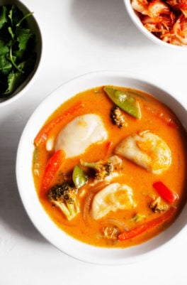 An overhead photograph of a warm bowl of red curry dumpling soup, with fresh herbs and kimchi on the side.