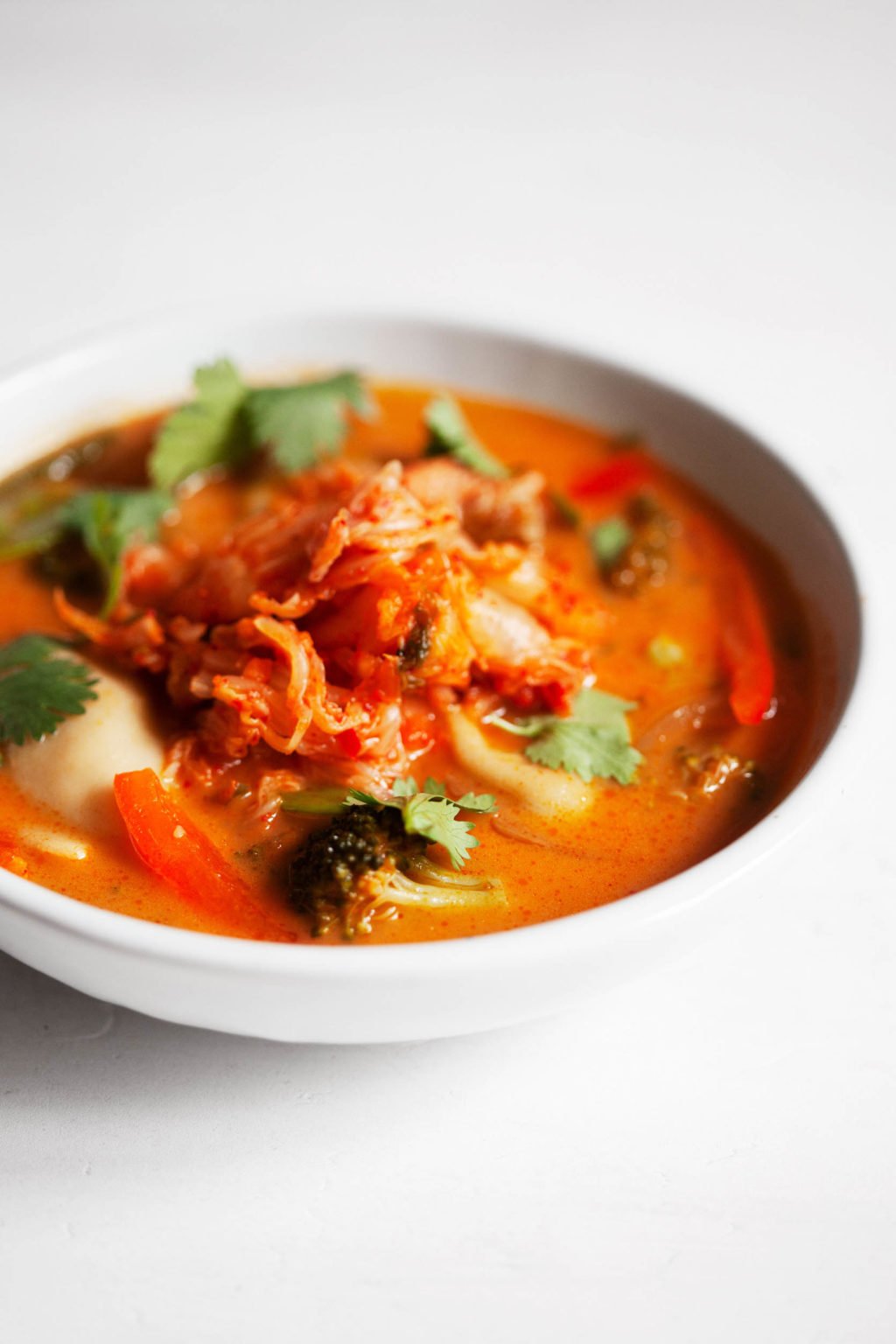 An angled photograph of a vegan red curry soup, made with plump, plant-based dumplings and garnished with a mound of kimchi.