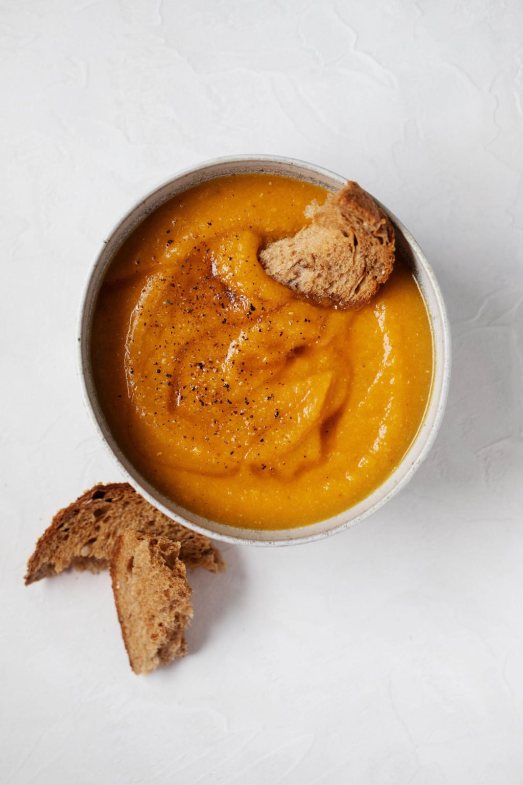 A plant based bowl of creamy, golden colored soup, with a small slice of bread dipped in for serving.