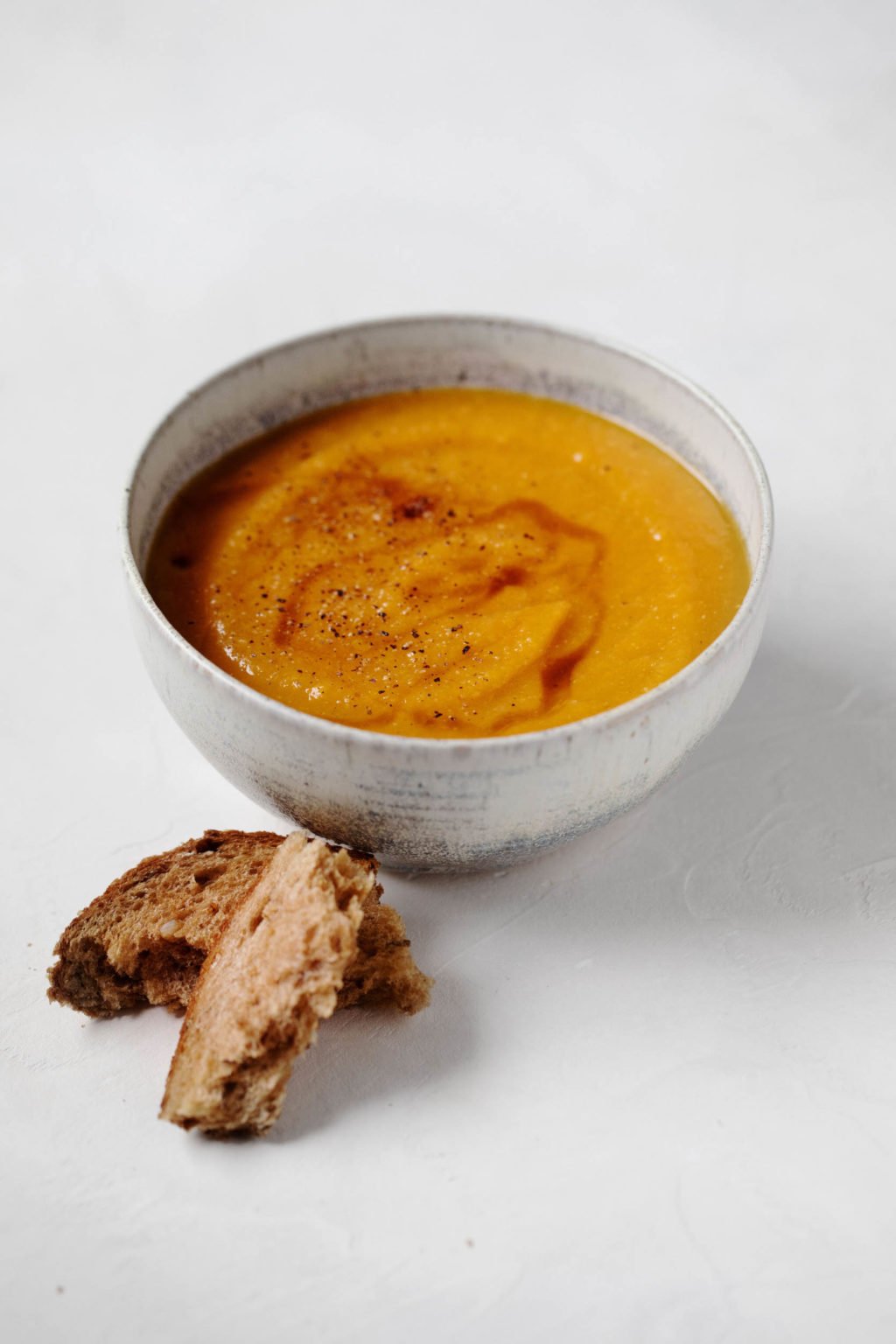 A bowl of vegan butternut lentil apple soup, garnished with a swirl of oil, freshly ground pepper, and a side of fresh bread.