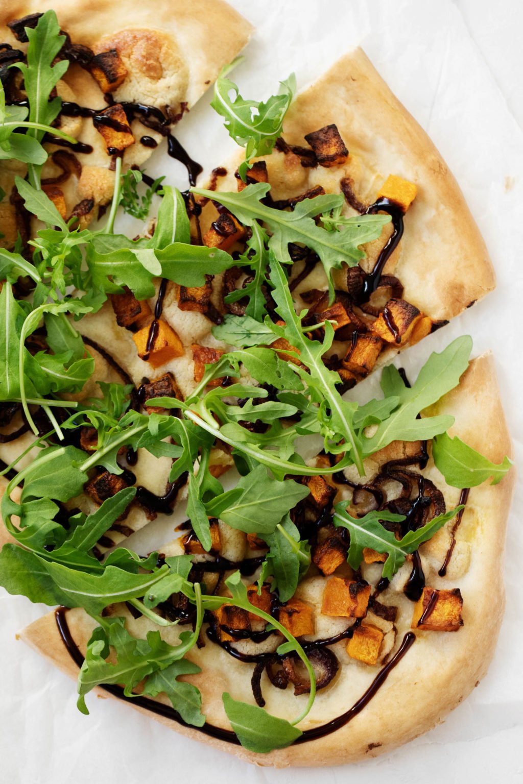 A vibrant and colorful vegan pizza for fall, topped with roasted cubes of butternuts squash, red onion, arugula, and a drizzle of balsamic glaze.