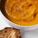 A small bowl of golden curried butternut squash and lentil soup is topped with a swirl of olive oil and accompanied by fresh bread.