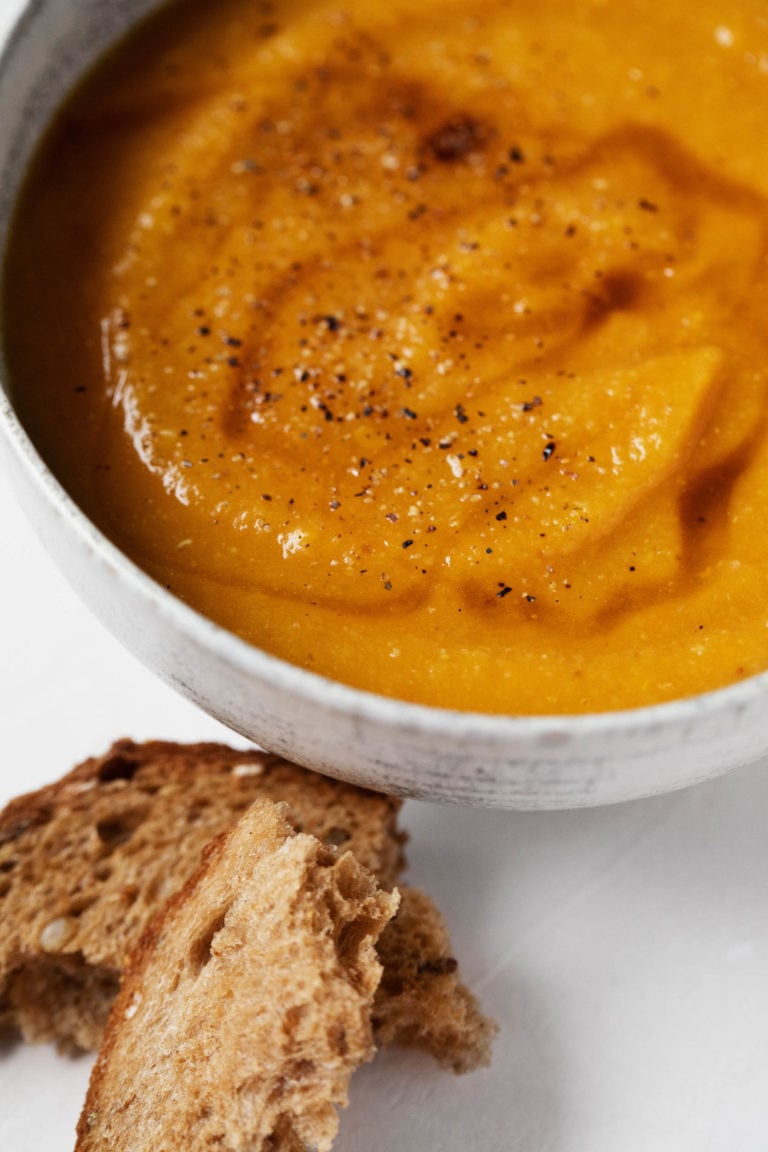 A small bowl of golden curried butternut squash and lentil soup is topped with a swirl of olive oil and accompanied by fresh bread.