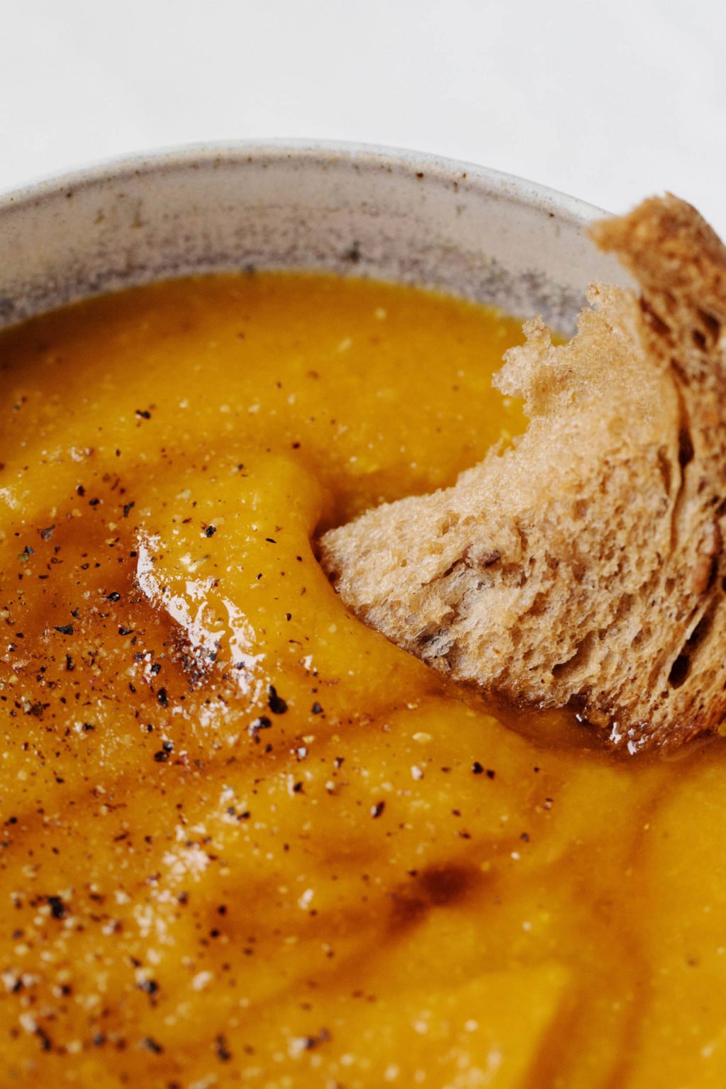A slice of whole grain bread is dunked into a bowl of creamy, orange colored vegan soup.