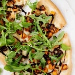 An overhead photo of vegan butternut red onion pizza, garnished generously with fresh arugula and drizzled with balsamic vinegar.