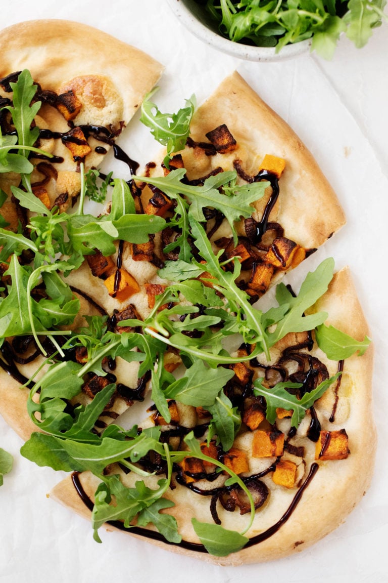 An overhead photo of vegan butternut red onion pizza, garnished generously with fresh arugula and drizzled with balsamic vinegar.