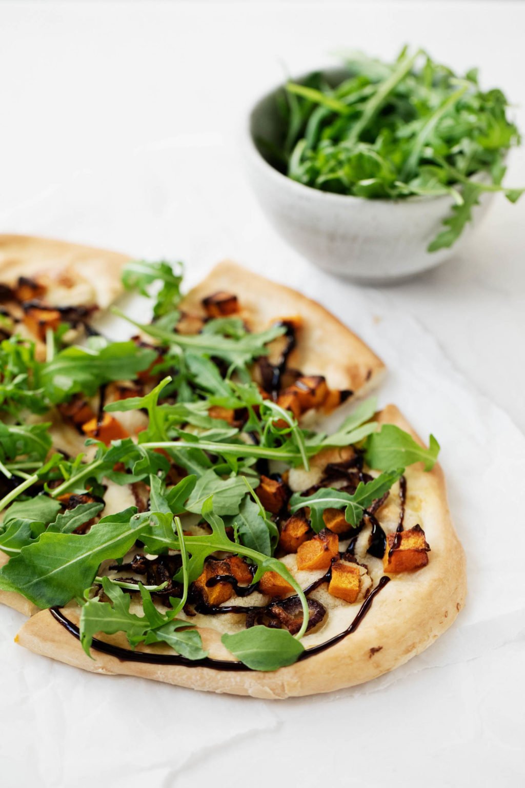 A vegan pizza with roasted butternut squash, red onions, and cashew cheese, topped with bright green arugula and drizzled with balsamic vinegar.
