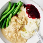 A bowl of vegan holiday food, including mashed potatoes, tempeh and gravy, green beans, and a little scoop of cranberry sauce.