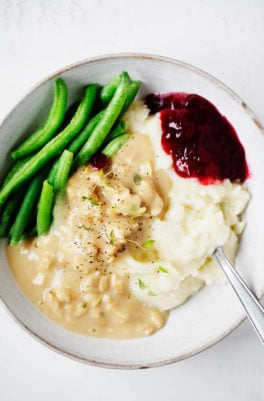 A bowl of vegan holiday food, including mashed potatoes, tempeh and gravy, green beans, and a little scoop of cranberry sauce.